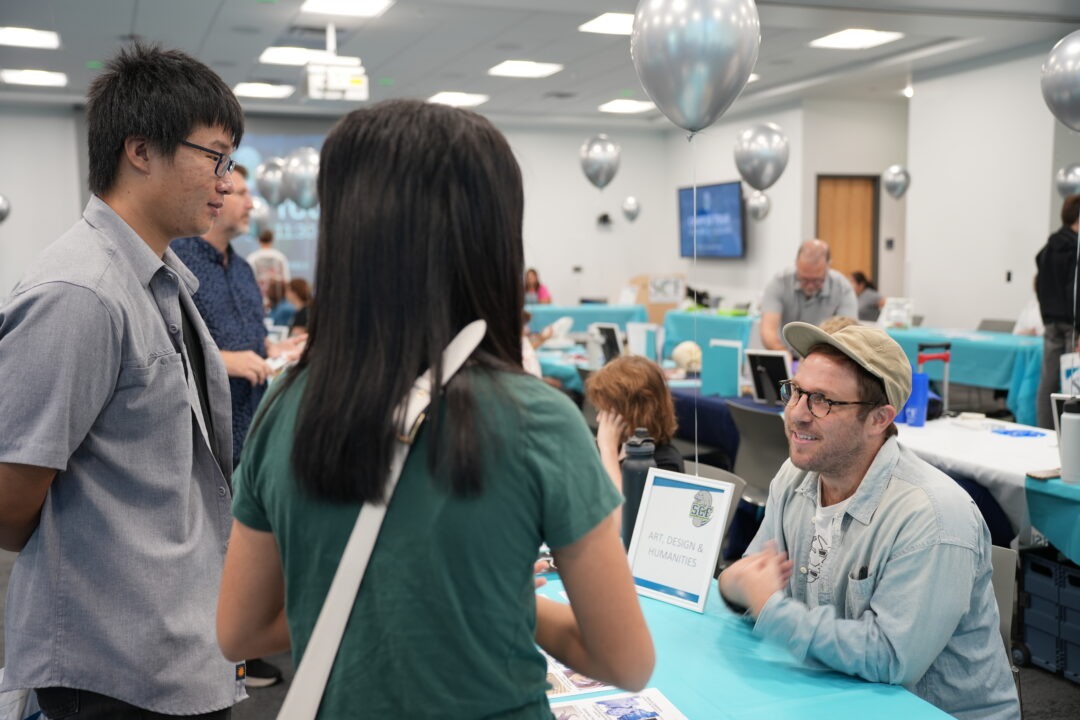 Two students talking a person in a hat sitting at the Art, Design and Humanities table during an open house event.